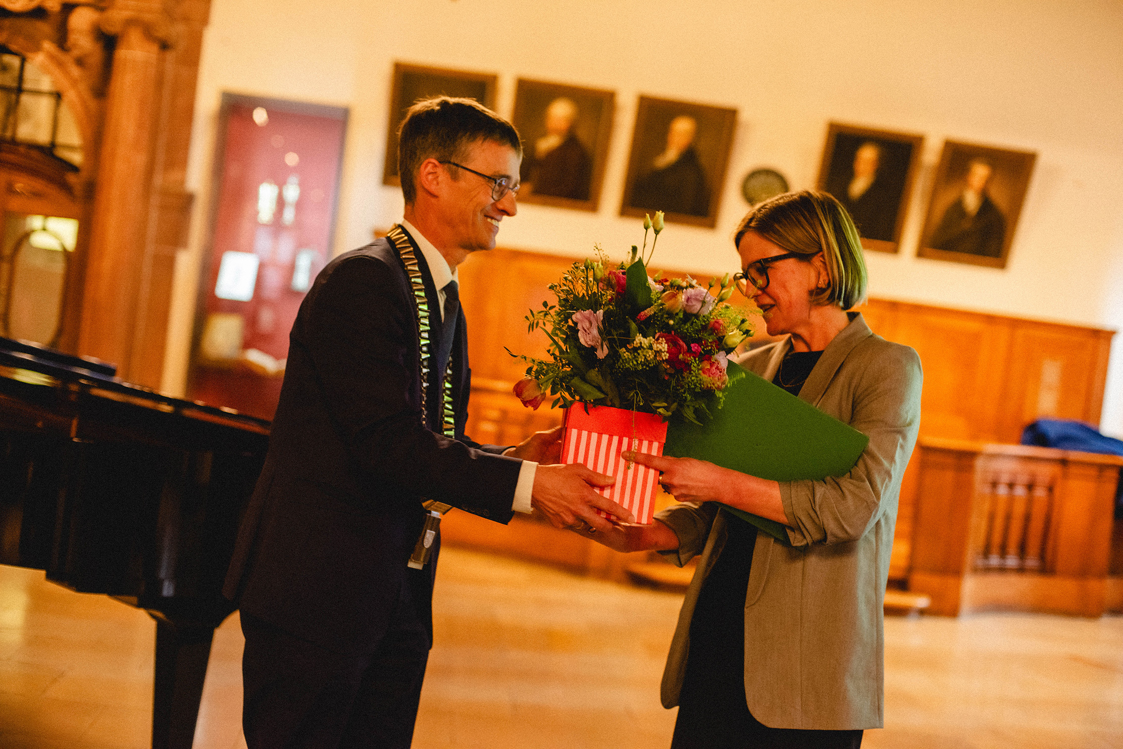 Eine männliche Person mit goldener Amtskette übergibt einer weiblichen Person mit Brille einen Blumenstrauß und eine grüne Urkunde. Im Hintergrund des Saales hängen Gemälde.