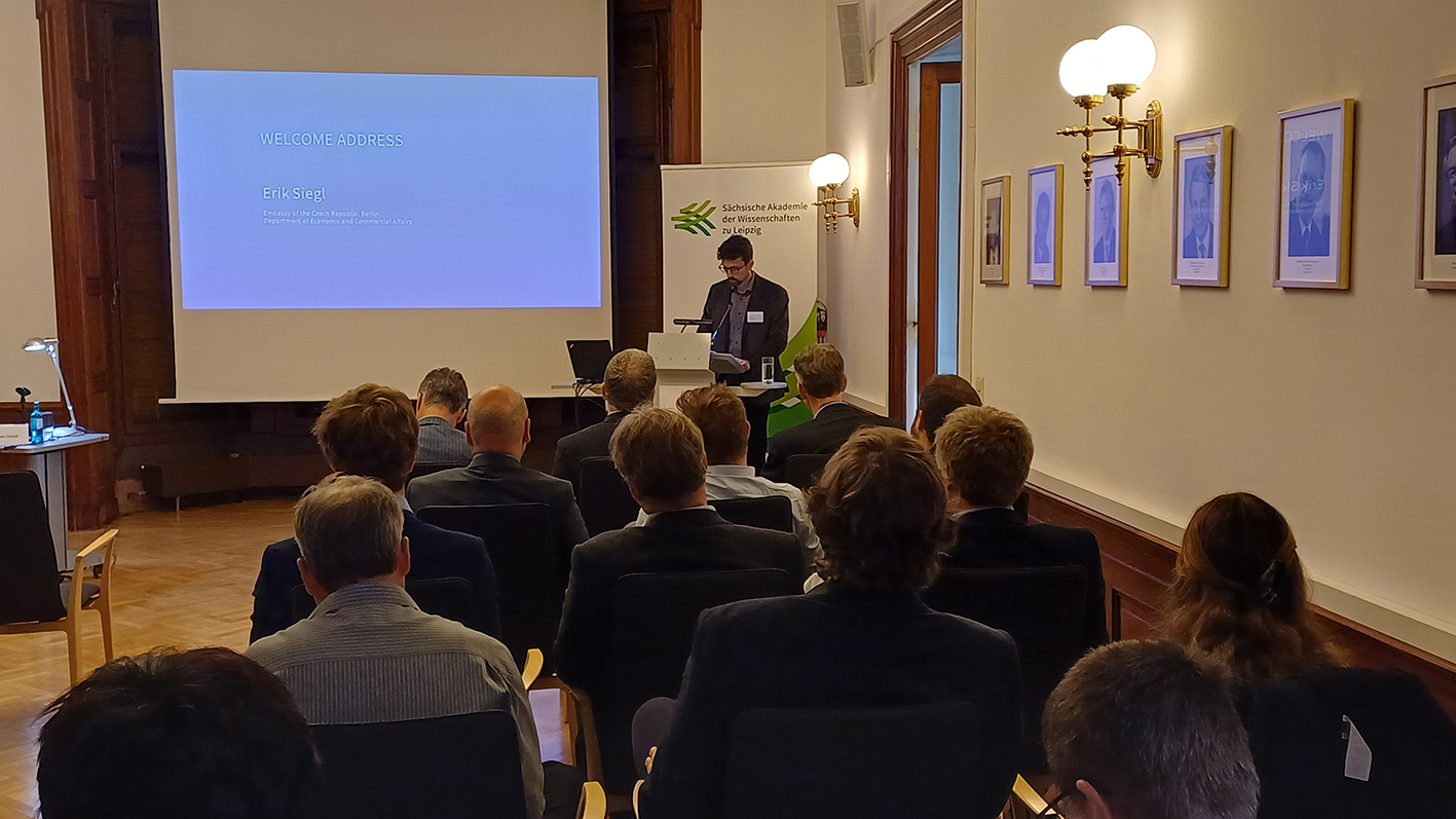 Eine männliche Person steht an einem Rednerpult und spricht. Mehrere andere Personen sitzen auf Stühlen und hören zu. Auf einer Projektions-Leinwand kann man auf blauem Hintergrund lesen "Welcome Address Erik Siegl". Hinter der vortragenden Person steht ein Aufsteller mit dem Logo der Sächsischen Akademie der Wissenschaften zu Leipzig.