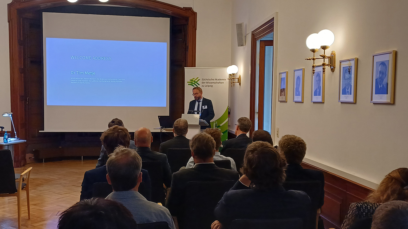 Eine männliche Person steht an einem Rednerpult und spricht. Mehrere andere Personen sitzen auf Stühlen und hören zu. Auf einer Projektions-Leinwand kann man auf blauem Hintergrund lesen "Welcome Address Dr Tim Metje". Hinter der vortragenden Person steht ein Aufsteller mit dem Logo der Sächsischen Akademie der Wissenschaften zu Leipzig.