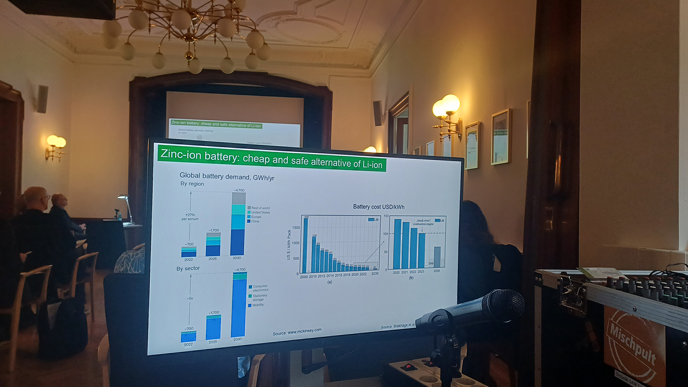 In einem Saal steht ein Bildschirm, auf dem man verschiedene Verteilungskurven erkennen kann. Die Überschrift darüber lautet "Zinc-ion battery: cheap and safe alternative of Li-ion". Hinter dem Bildschirm sitzen Personen auf Stühlen und verfolgen einen Vortrag.