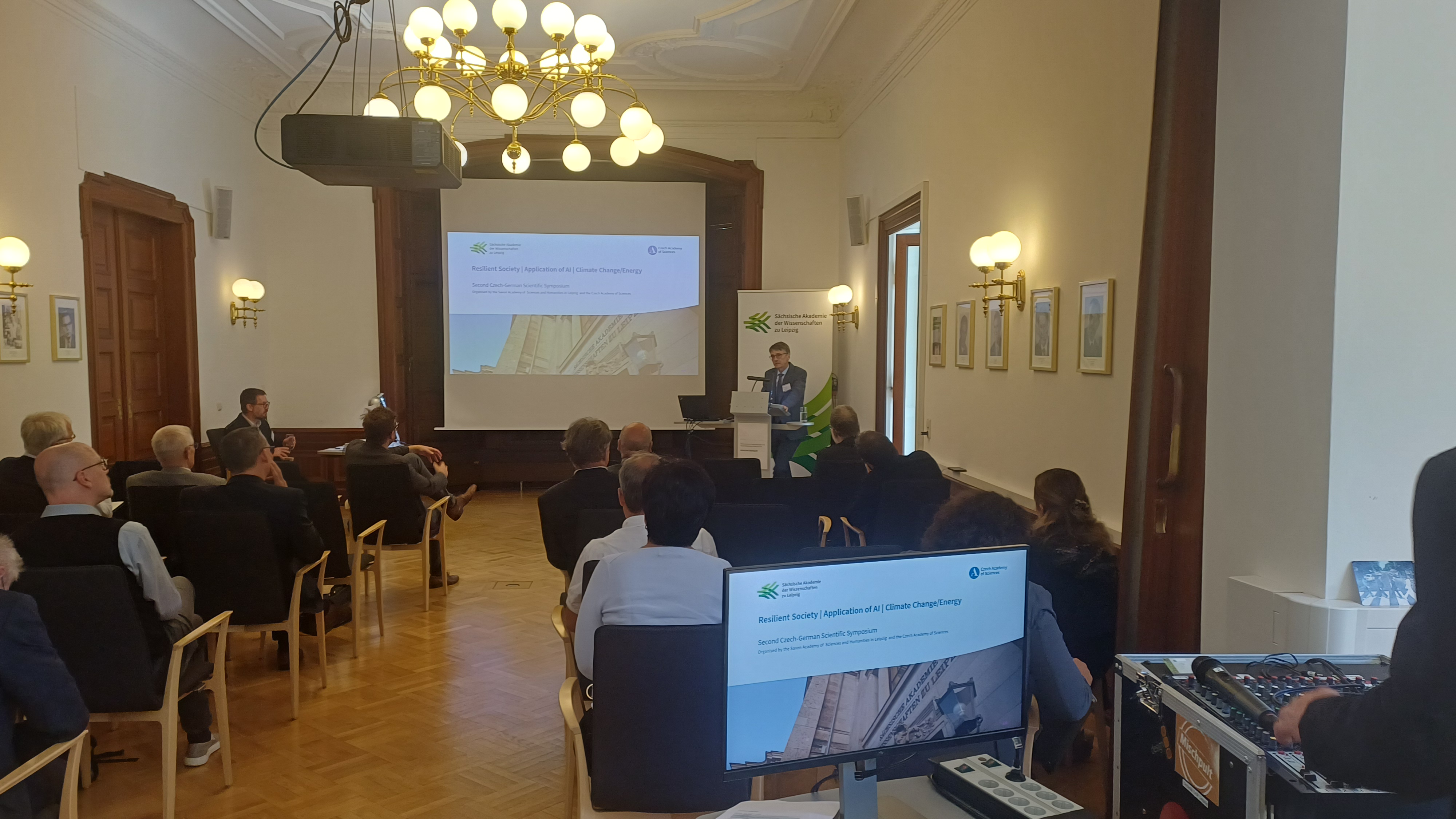 Eine männliche Person steht an einem Rednerpult und spricht. Mehrere andere Personen sitzen auf Stühlen und verfolgen den Vortrag. Auf einer Projektions-Leinwand kann man lesen "Resilient Society, Application of AI, Climate Change/Energy", darüber finden sich die Logos der Sächsischen und der Tschechischen Akademie der Wissenschaften. Hinter der vortragenden Person steht ein Aufsteller mit dem Logo der Sächsischen Akademie der Wissenschaften zu Leipzig.