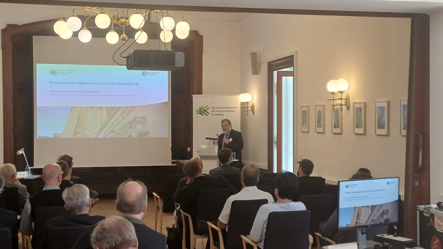 Eine männliche Person steht an einem Rednerpult und spricht. Mehrere andere Personen sitzen auf Stühlen und verfolgen den Vortrag. Auf einer Projektions-Leinwand kann man lesen "Resilient Society, Application of AI, Climate Change/Energy", darüber finden sich die Logos der Sächsischen und der Tschechischen Akademie der Wissenschaften. Hinter der vortragenden Person steht ein Aufsteller mit dem Logo der Sächsischen Akademie der Wissenschaften zu Leipzig.