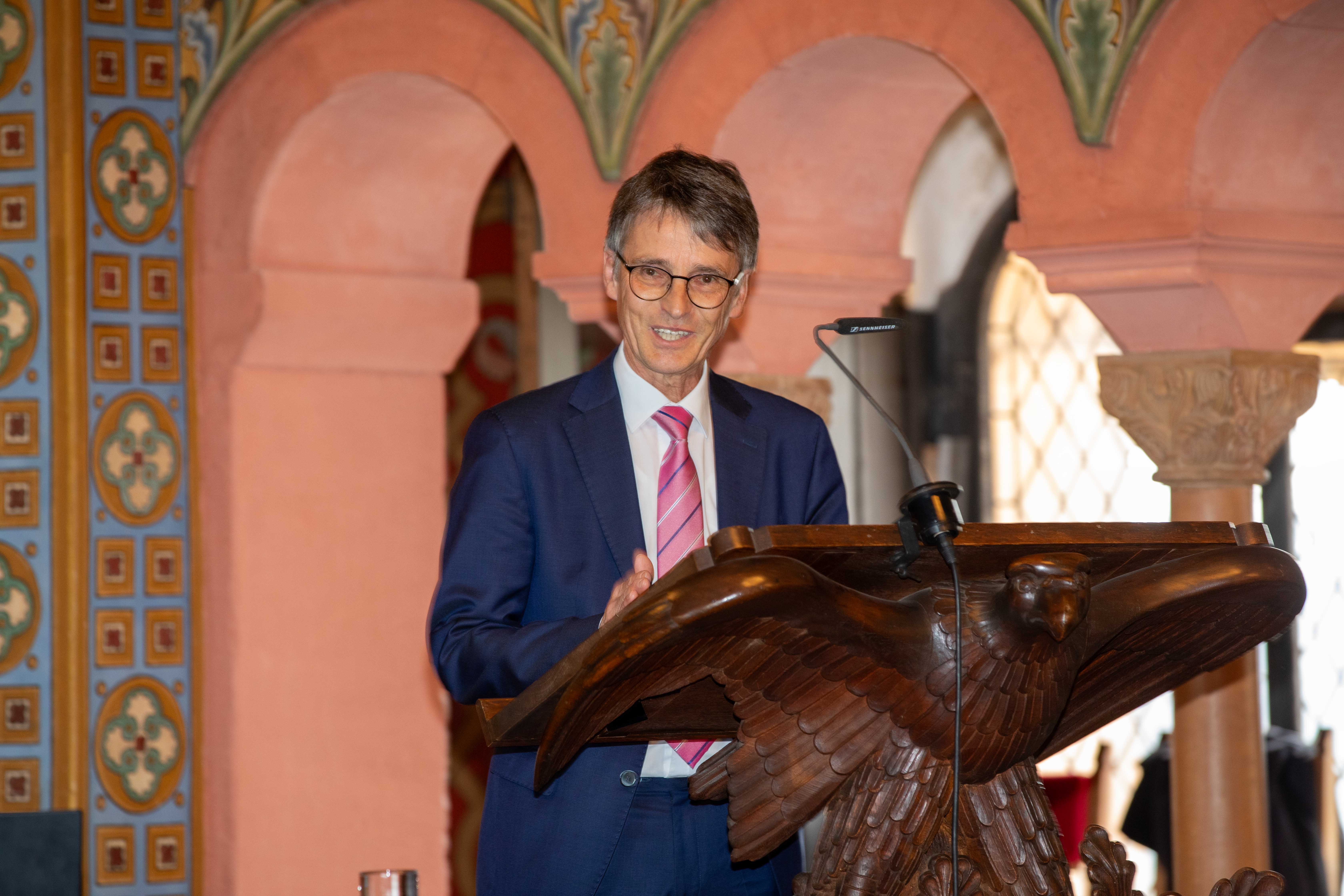 Eine männliche Person mit Brille steht in historischem Festsaal an einem Rednerpult, das von einem hölzernen Vogel getragen wird, und spricht in ein Mikrofon.