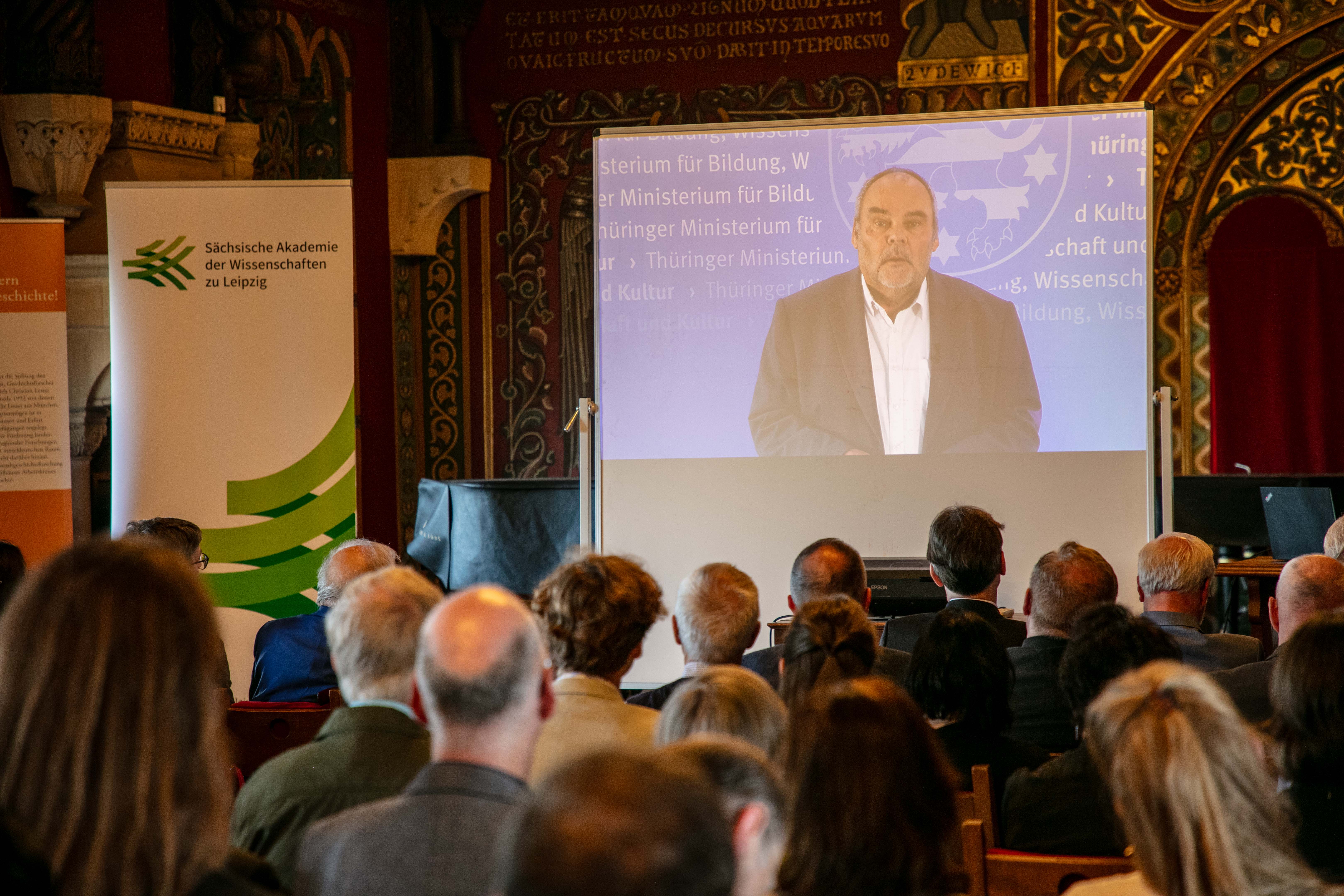 Auf einer Leinwand in einem historischen Festsaal wird ein Video abgespielt, in dem eine männliche Person vor blauem Hintergrund spricht. Im Vordergrund sitzen Personen auf Stühlen. Links sieht man einen Aufsteller mit dem Logo der Sächsischen Akademie der Wissenschaften zu Leipzig.