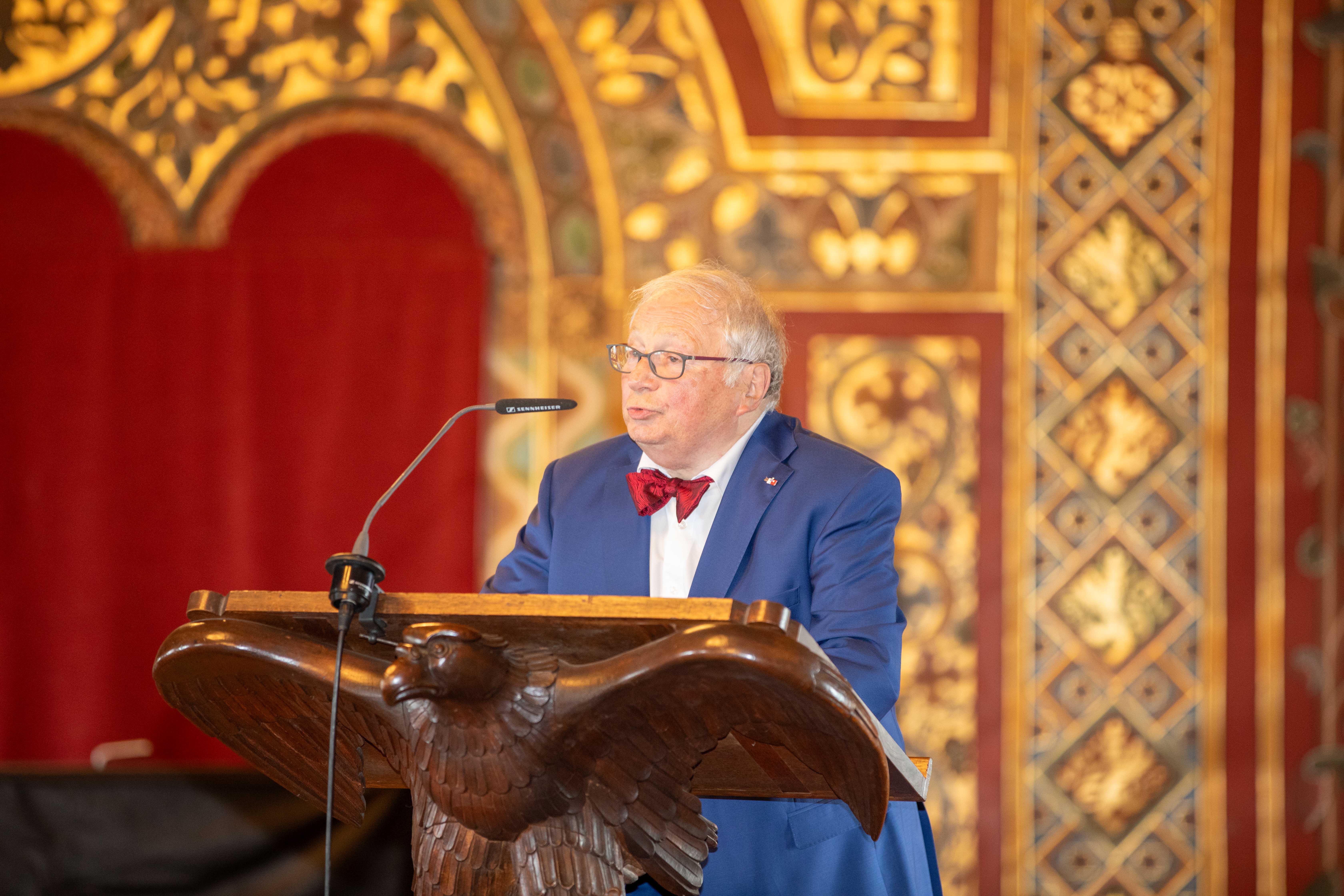 Eine männliche Person mit Brille steht in einem historischen Festsaal an einem Rednerpult, das von einem hölzernen Vogel getragen wird, und spricht in ein Mikrofon.