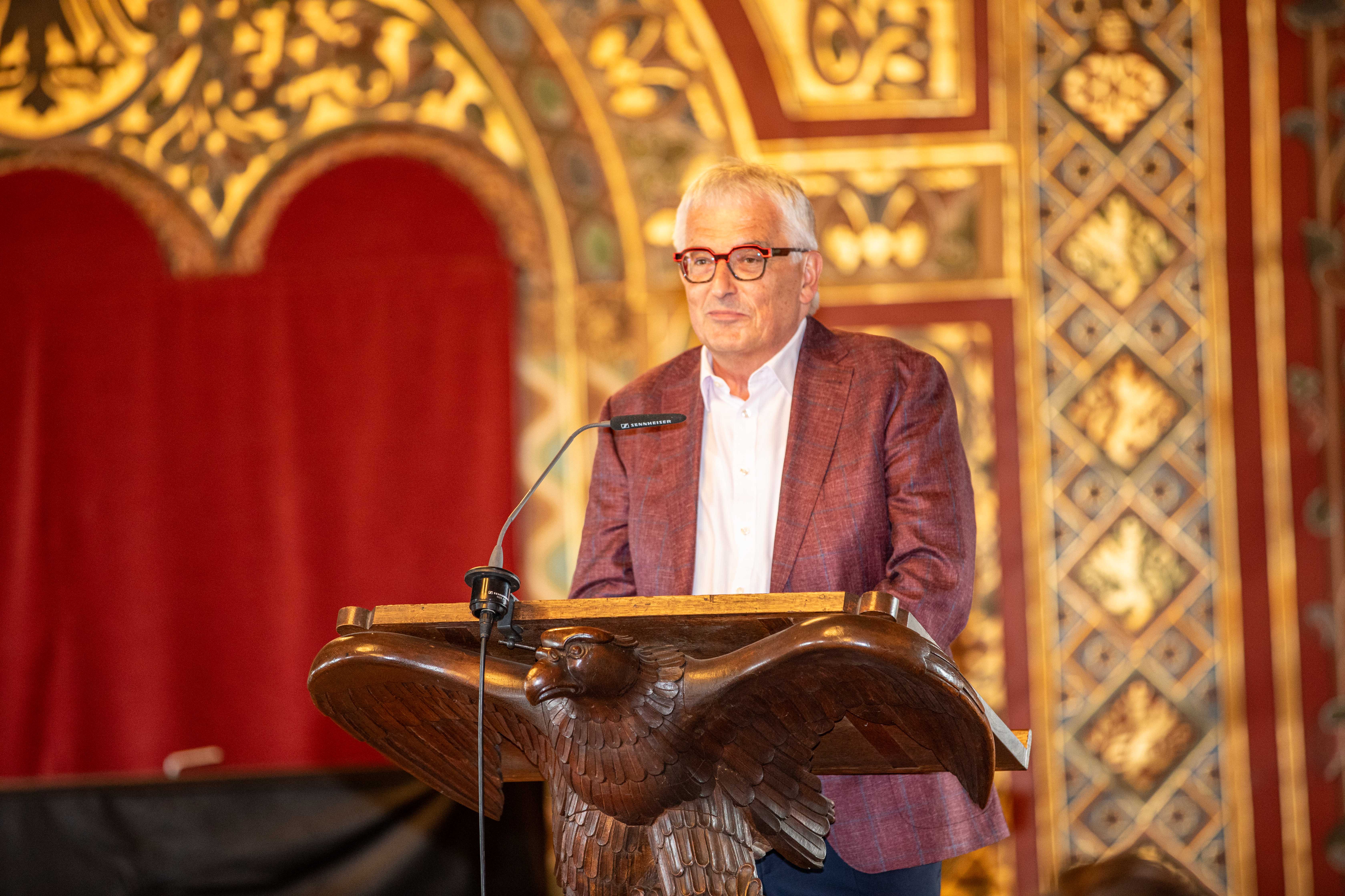 Eine männliche Person mit Brille steht in einem historischen Festsaal an einem Rednerpult, das von einem hölzernen Vogel getragen wird, und spricht in ein Mikrofon.