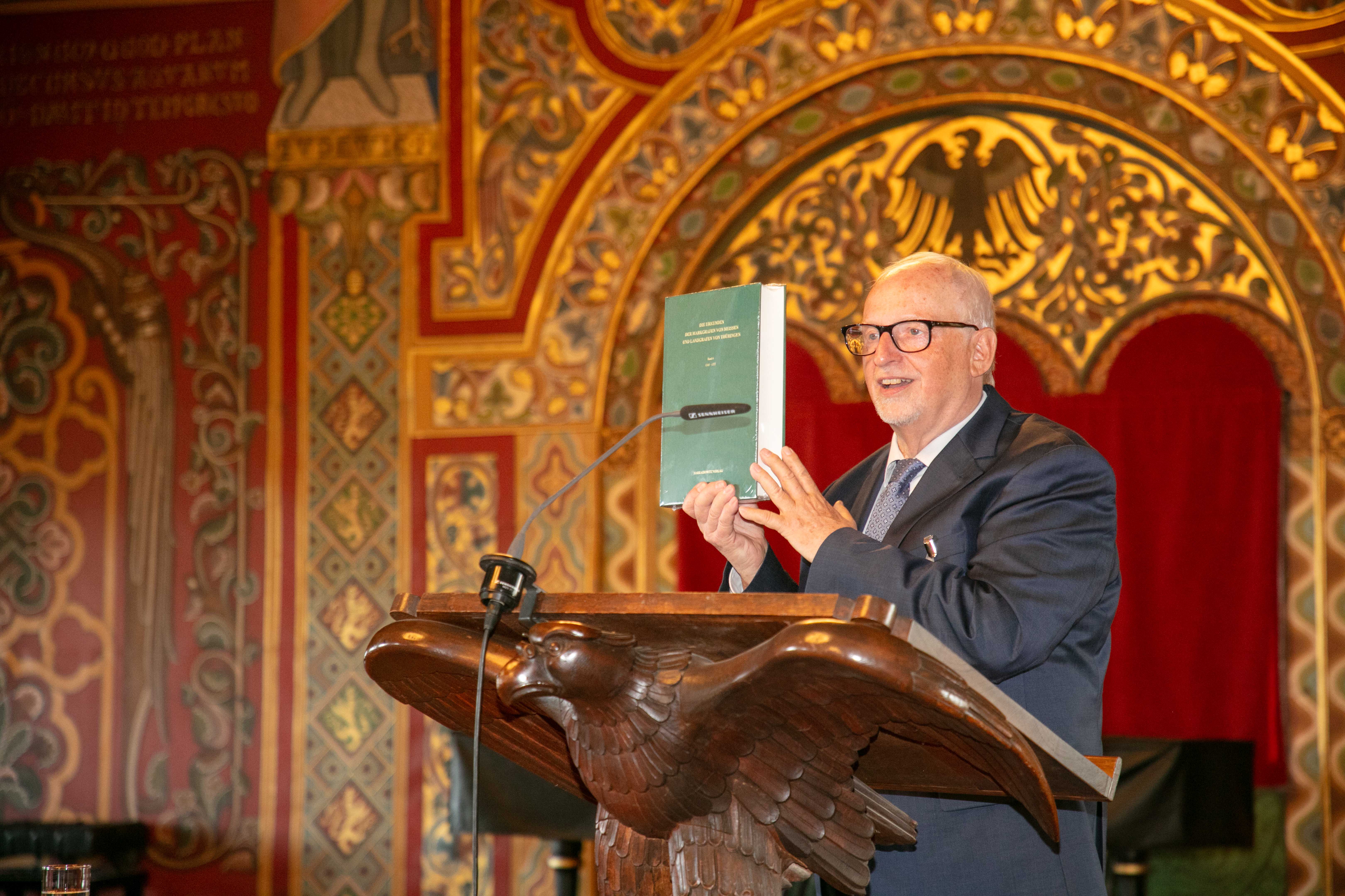 Eine männliche Person mit Brille steht in einem historischen Festsaal an einem Rednerpult, das von einem hölzernen Vogel getragen wird, und hält ein grünes Buch in den Händen.