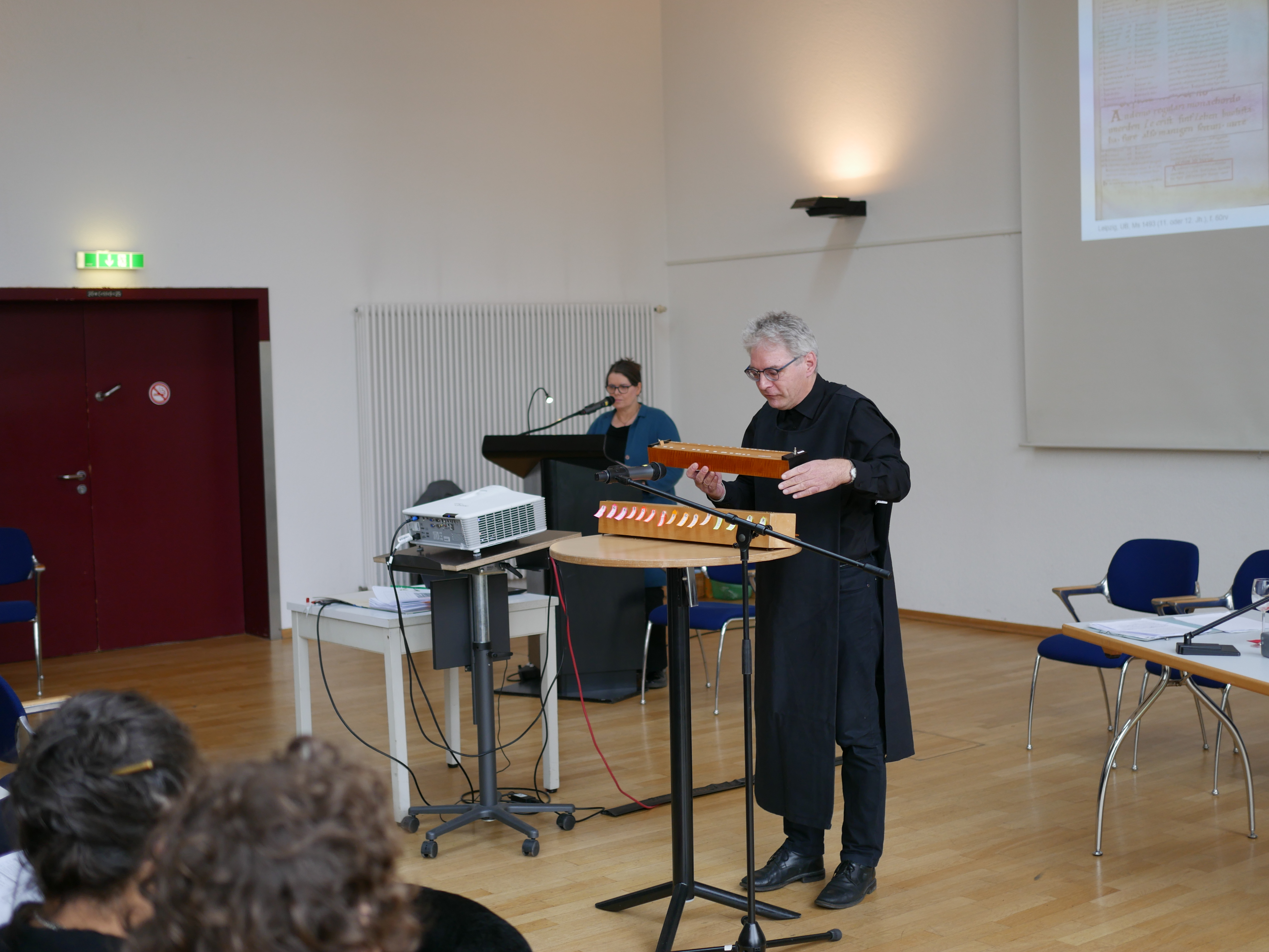 Eine Frau (links) erläutert etwas am Pult im Hintergrund. Ein Mann (rechts) demonstriert in schwarzer historischer Kleidung, wie ein Monochord, das ist ein Musikinstrument, funktioniert. Dieses sieht aus wie ein länglicher Holzkasten. An einer Wand ist ein Teil der Präsenation zu sehen.