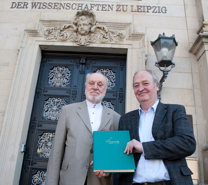 Vor der Eingangstür der Sächsischen Akademie der Wissenschaften stehen zwei männliche Personen. Einer hält ein grünes Buch in der Hand.