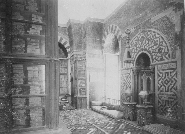 Black-and-white photograph of an old library with ornate arches, bookshelves and cushions on the mosaic floors